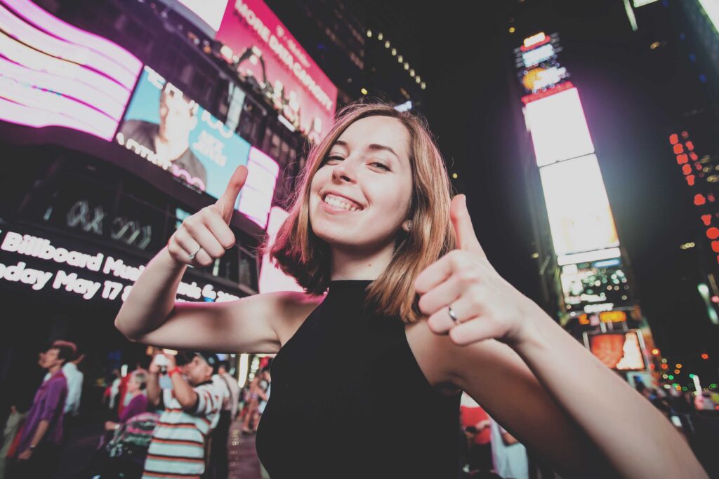 Junge Frau in New York am Times Square lächelt mit Daumen nach oben in die Kamera.